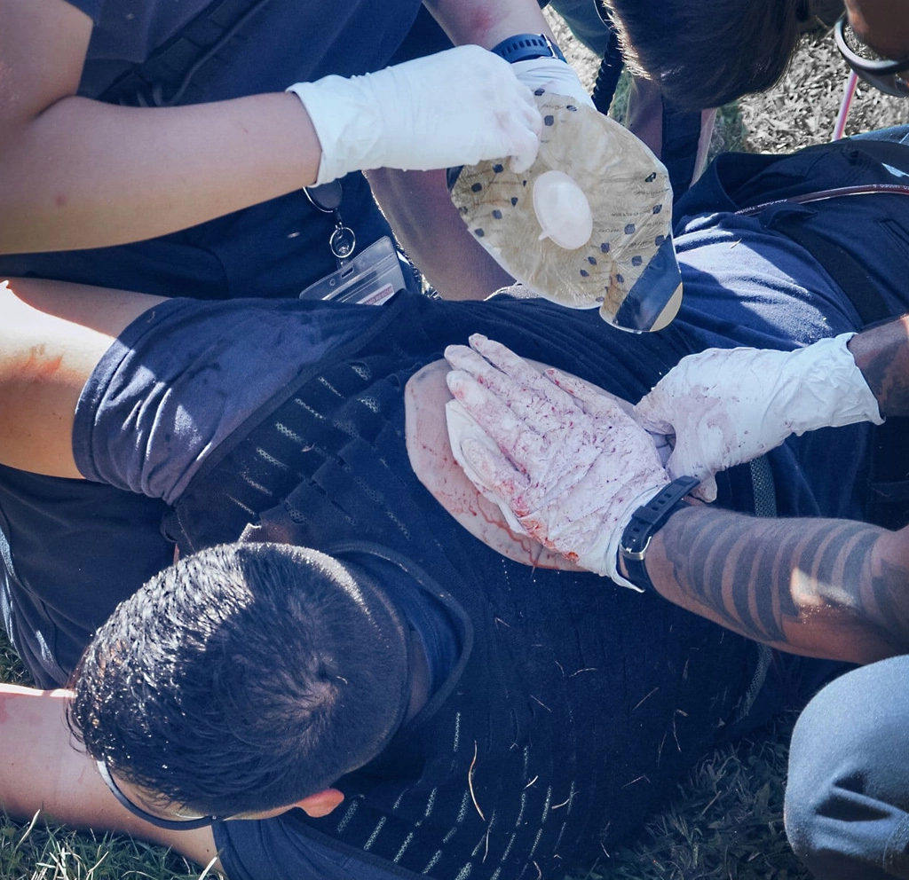 First aid trainees treating a simulated chest wound on a casualty wearing a trauma training vest during a hands-on emergency scenario.
